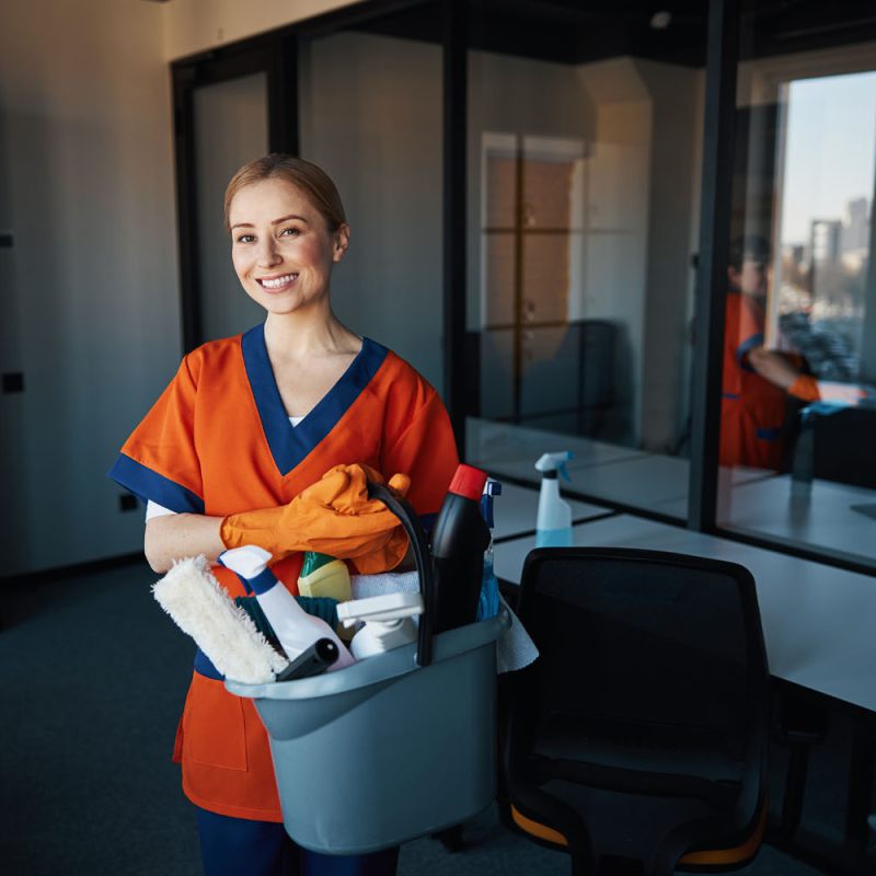 Reinigungskraft mit orangenem Shirt und Putzeimer lächelt in die Kamera.